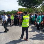 A group of people gathered outdoors, some wearing safety vests. A man in a green vest walks in the foreground.