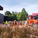 Fire truck and emergency personnel near a building and tall grass on a sunny day. Responders wear orange gear.