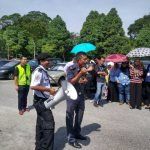 Two security guards with a megaphone addressing a crowd outdoors on a sunny day, some holding umbrellas.