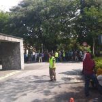 A group of people gathered outside. A person in a yellow vest stands in the foreground. Trees and a building are in the background.