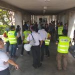 A group of people, many wearing yellow vests, gathered in a covered outdoor walkway. A person is speaking into a megaphone.