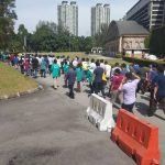A large group of people walking along a road, possibly an event or parade. Two tall buildings and a brick building are in the background.