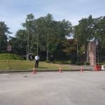 A person in a white shirt stands next to orange traffic cones on a paved area, with trees and a hill in the background.