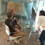 Three people talking in an office setting. One man sits in a chair; a police officer stands between him and another person facing them.