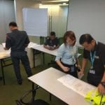 People in a conference room examine papers. Two men in dark uniforms, one in front and one in the background, and two colleagues in casual wear.