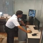 A man in a white shirt and hat operates a computer with a security camera feed. He is in an office setting, leaning towards the monitor.