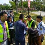 A group of people, some wearing safety vests, talking on a sidewalk near a park. A security guard stands nearby.