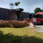Fire truck at a building, with firefighters in the background and a man walking toward it on a sunny day.