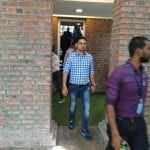 People walking through a doorway with brick walls. A man in a checkered shirt and jeans is in focus, with others blurred in the background.