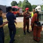 Firefighters in uniform stand near a fire truck, discussing details. The scene takes place outdoors with trees in the background.