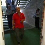 Man in red shirt walking toward the viewer; staircase and brick wall in the background. A person plays the trumpet.