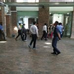 People walking in a hallway or entrance area with glass doors and brick pillars. Some are wearing white shirts and dark pants.