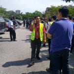 Man in yellow vest talking on phone in a parking lot, surrounded by people and cars. Sunlight casts shadows.