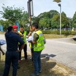 A group of men in safety vests and casual clothing stand outdoors, possibly discussing documents near a road.