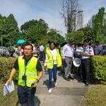 People in bright safety vests walk along a sidewalk near trees and a building on a sunny day.