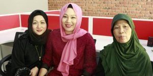Three women wearing hijabs pose together. One in black, one in pink, and one in green, smiling in an office setting.