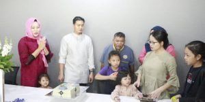 A group of people, including children, gather around a table, possibly celebrating. Two women in hijabs, one in red, stand near the front.