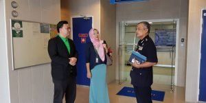 Three people in an office setting. A man in a black suit with a green shirt, a woman in a hijab and blue skirt, and a man in a dark uniform are talking.