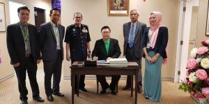 A group of people gathered indoors, with one person seated at a desk signing a document. Others stand around, some in suits, a uniformed officer, and a woman in a hijab.