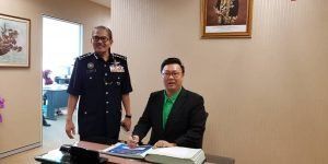A man in a black suit and green shirt sits at a desk signing a document, a uniformed officer stands smiling behind him. They are in an office setting.
