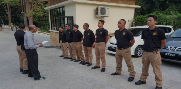 A group of security personnel in uniform standing in line while being addressed by a man holding papers near a building and vehicles.