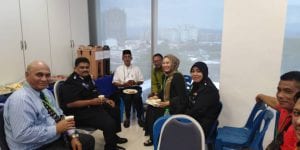 A group of people indoors, some seated and others standing, eating and drinking in a room with large windows overlooking a cityscape.