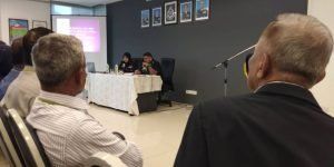 People in a conference room, with presenters at a table and audience members in the foreground. A presentation is on a screen.