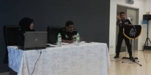 A presentation with three people at a table and one person speaking into a microphone, inside a conference room.