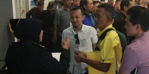 A woman in a black uniform talks to a group of men in a crowded setting. One man wears a yellow shirt.