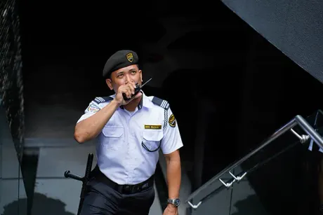 Security guard in white uniform and blue beret walks down stairs, holding a walkie-talkie.