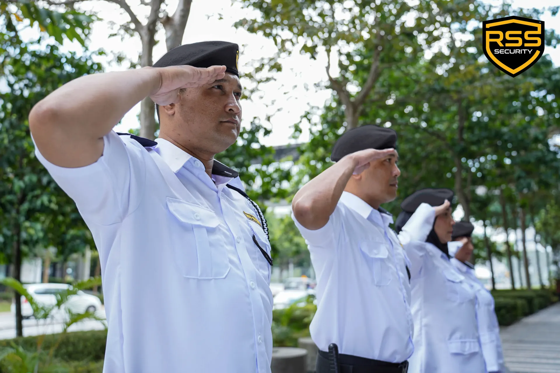 Uniformed people in black berets saluting outdoors near trees and a building