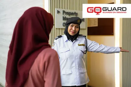 A security guard in a blue beret and uniform guides a person wearing a maroon hijab. The guard is in an office doorway.