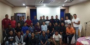 A group of people poses for a photo in a conference room. Most are men, wearing various casual attire. A few people stand, the rest sit.