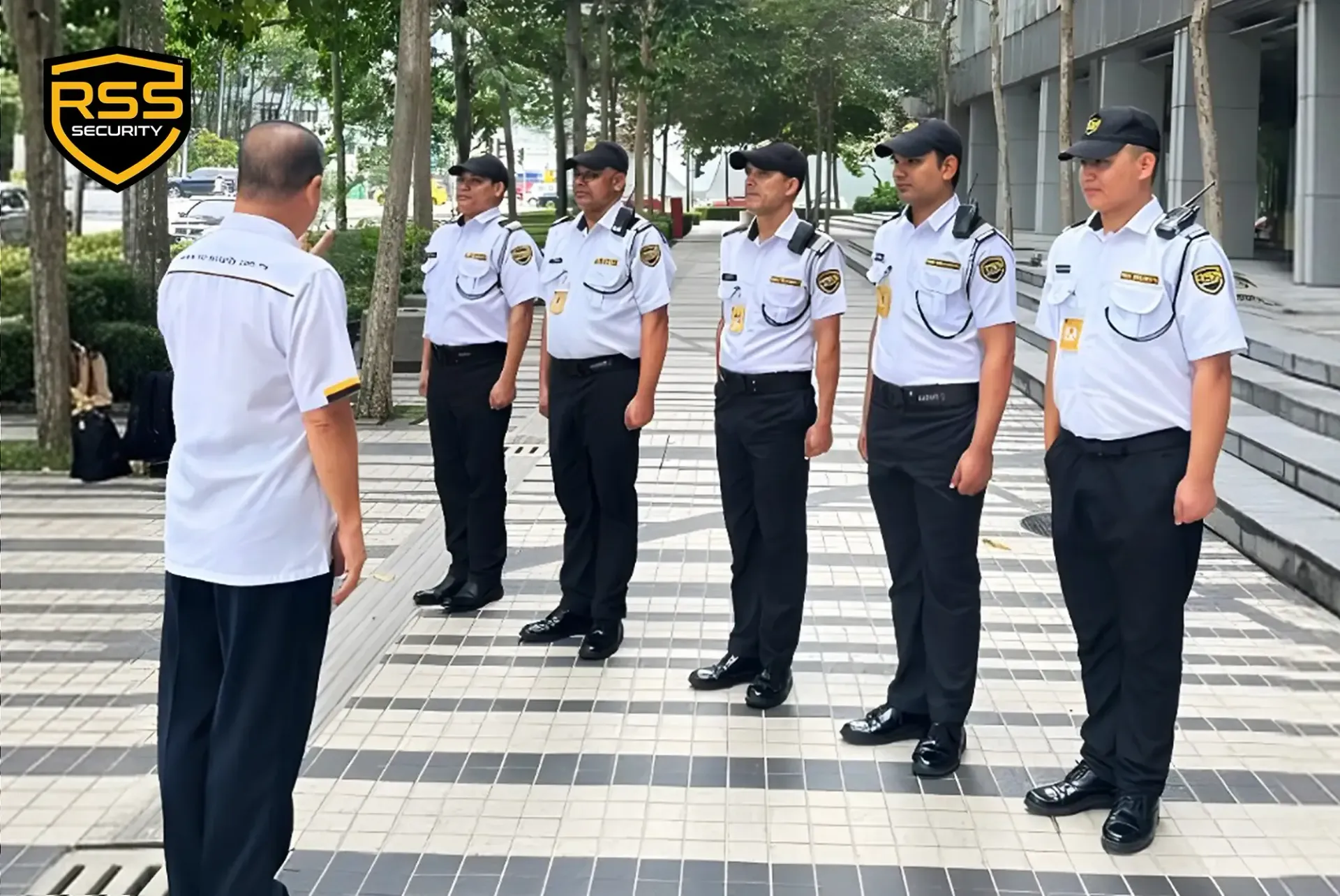 A supervisor in a black uniform briefing four security personnel in white shirts and black trousers in an outdoor lot.
