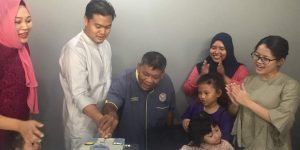 A group of people celebrates with a cake cutting. A man in a blue uniform cuts the cake while others clap and smile, likely at a birthday or other celebration.