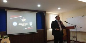 A man in a suit speaks at a podium during a presentation, with a screen displaying a logo behind him. The room has blue walls and whiteboards.