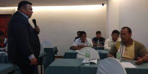 A man in a suit speaks into a microphone at a conference table. Several men sit at the table, listening.