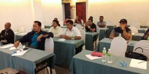 A group of people are seated at tables in a conference room, appearing to be attending a training or meeting.