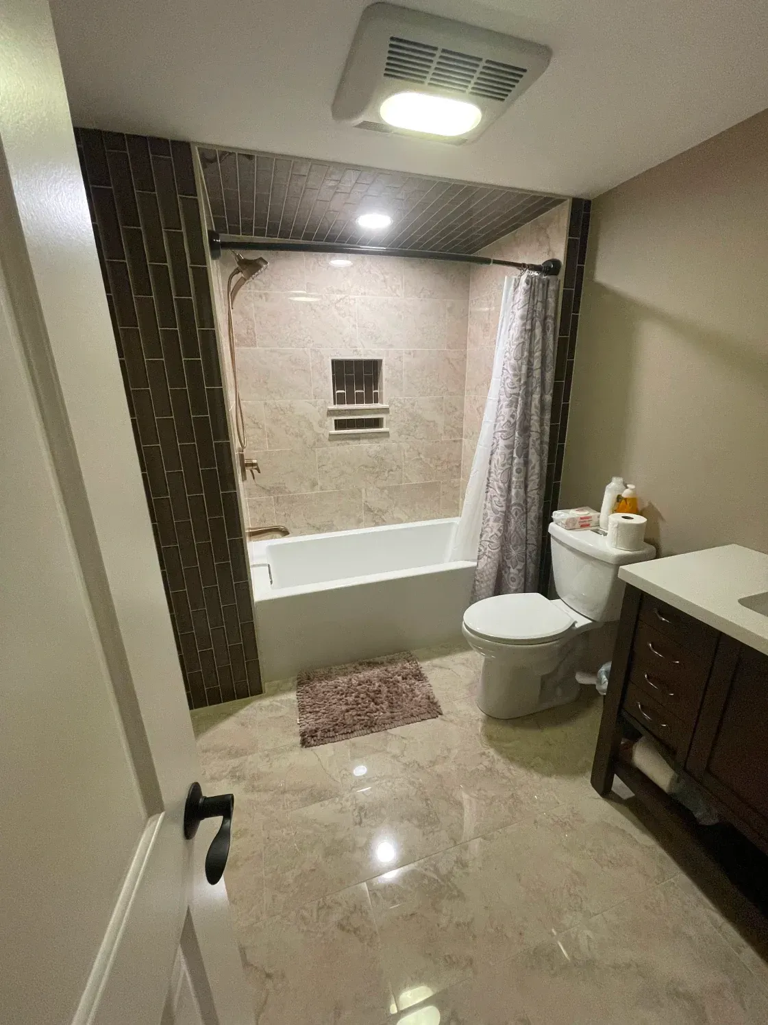 Bathroom with a white tub, toilet, and vanity. Brown accent wall, tan tile, and patterned shower curtain.