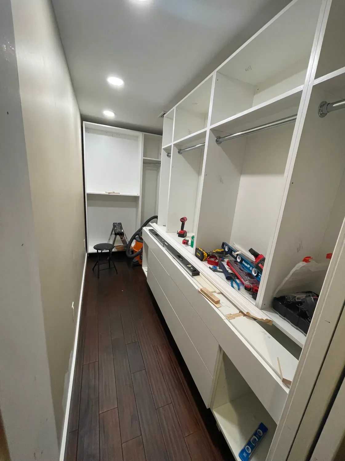 Walk-in closet under construction with white shelving, a long dark wood floor, and tools scattered about.