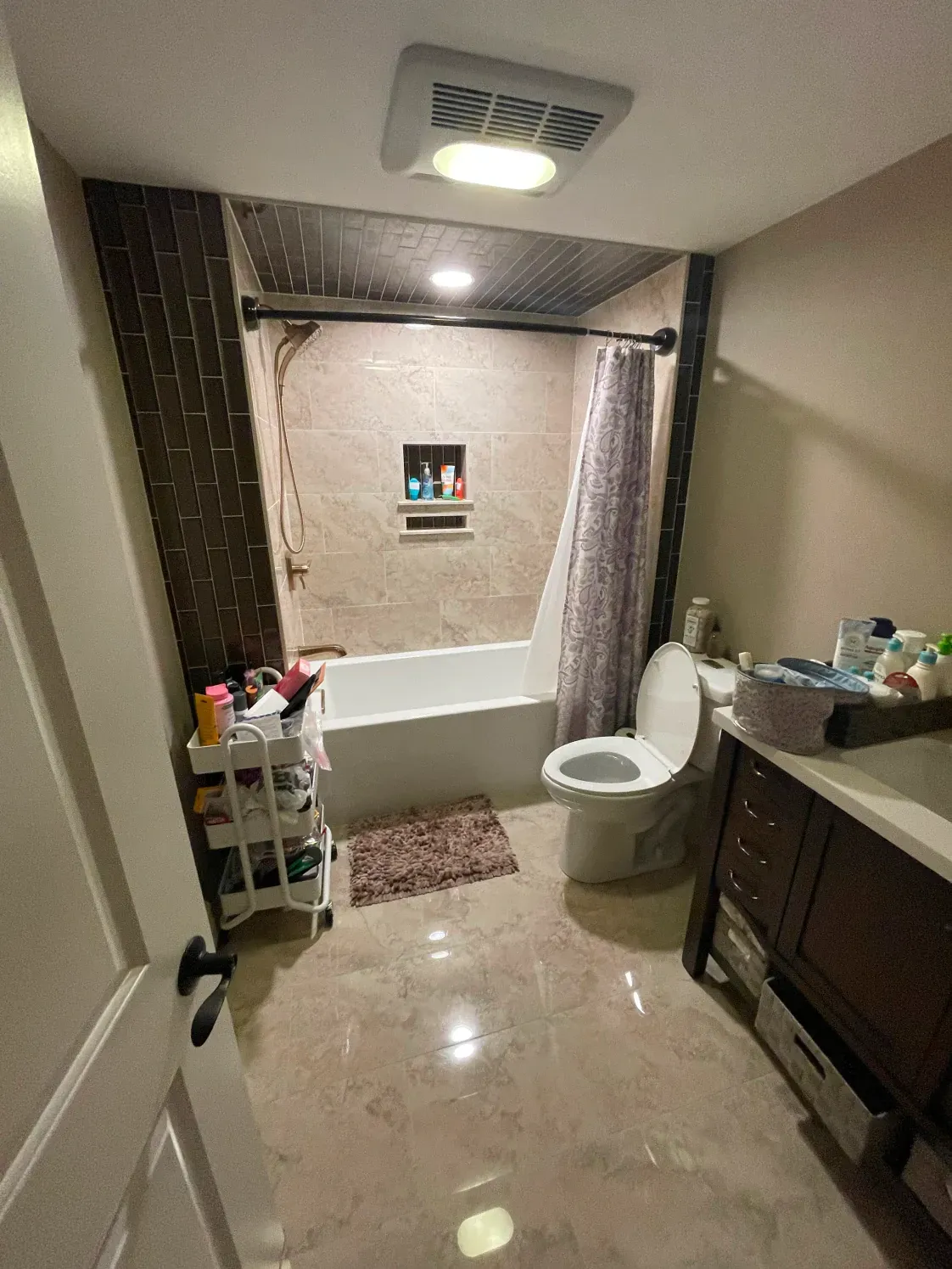 Bathroom with tub/shower, toilet, vanity, and small rolling storage cart. Neutral tones.