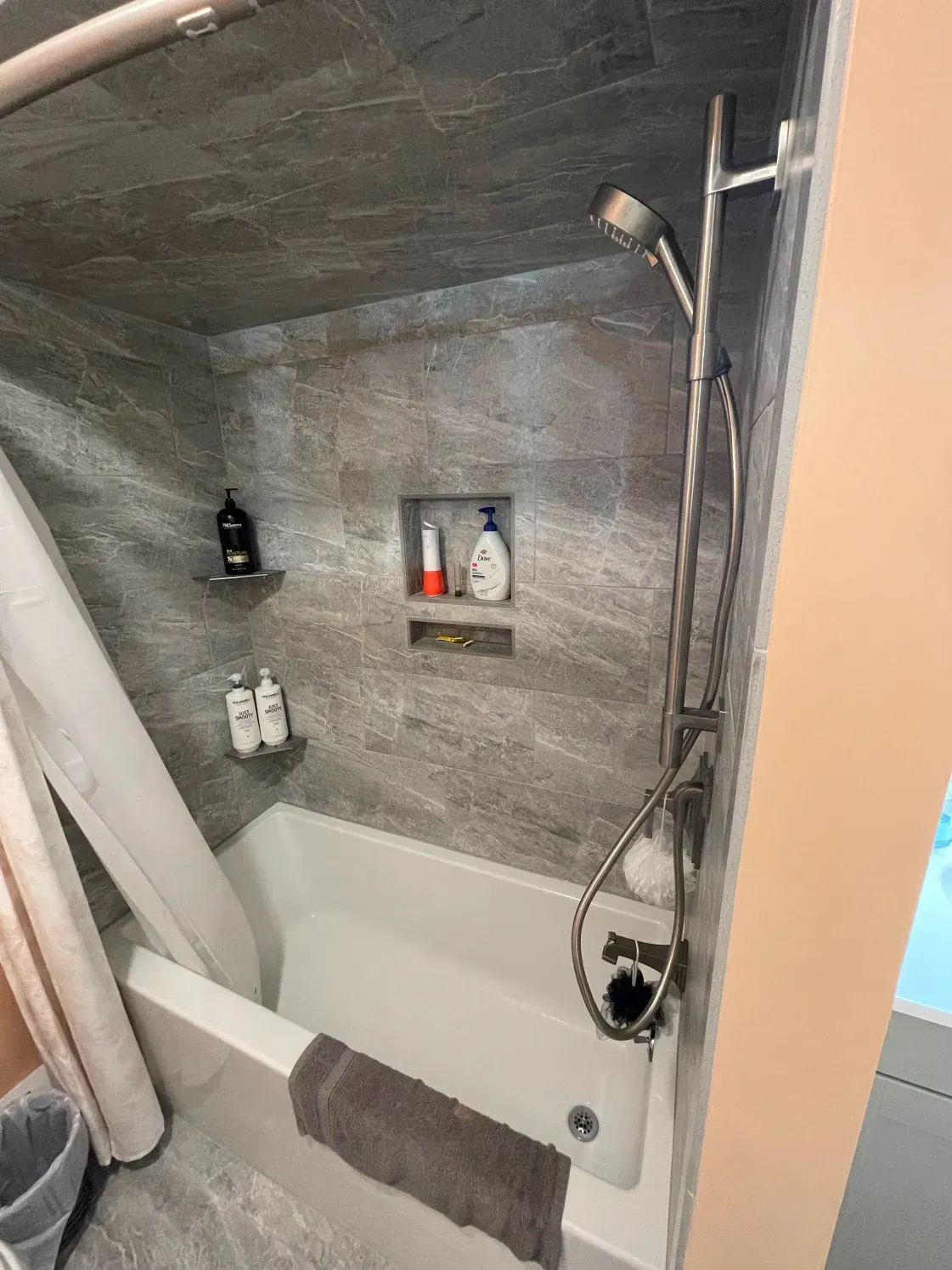 Bathroom with gray tile walls, a bathtub, showerhead, and a built-in shelf with toiletries.