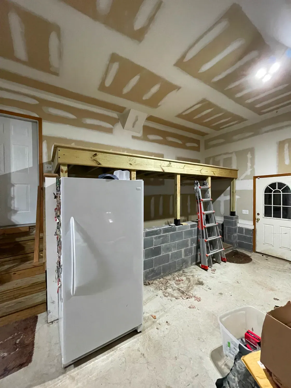 Interior of unfinished room, refrigerator, cinder block structure, ladder, exposed studs, drywall.