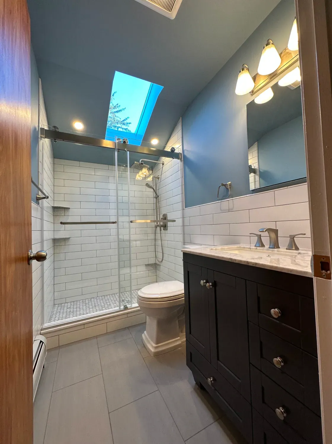 Bathroom with blue walls, skylight, dark vanity, white and grey tile, and a glass shower.