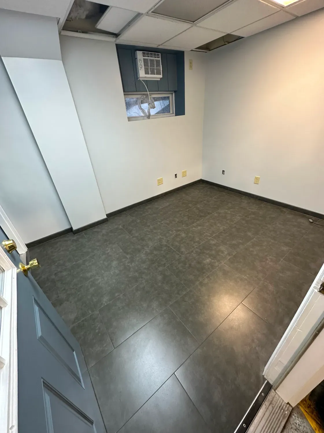 Empty office room, gray flooring, light blue walls, small window with AC unit, missing ceiling tiles, open blue door.