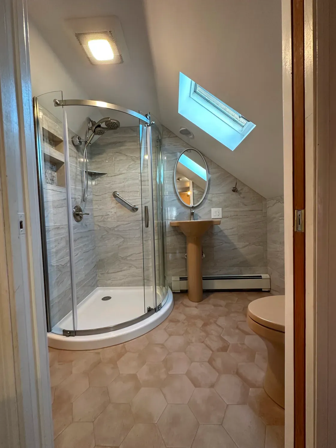 Bathroom with a curved glass shower, pedestal sink, skylight, and tan hexagonal floor tiles.