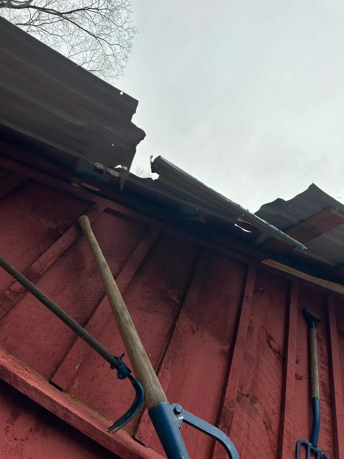Red wooden shed with damaged corrugated metal roof and tools hanging on the side.