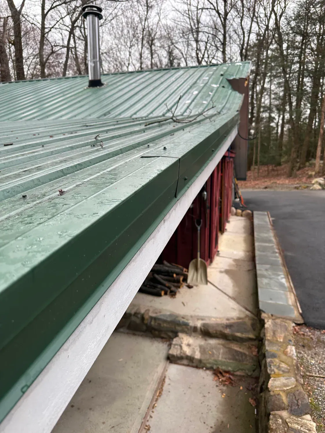 Green metal roof overhangs a red building with wood pile and stone steps.