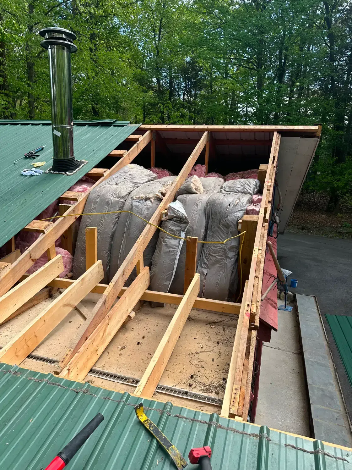 Roof partially open, revealing rafters and large insulation bags. Green roof, metal chimney, and wooded background.