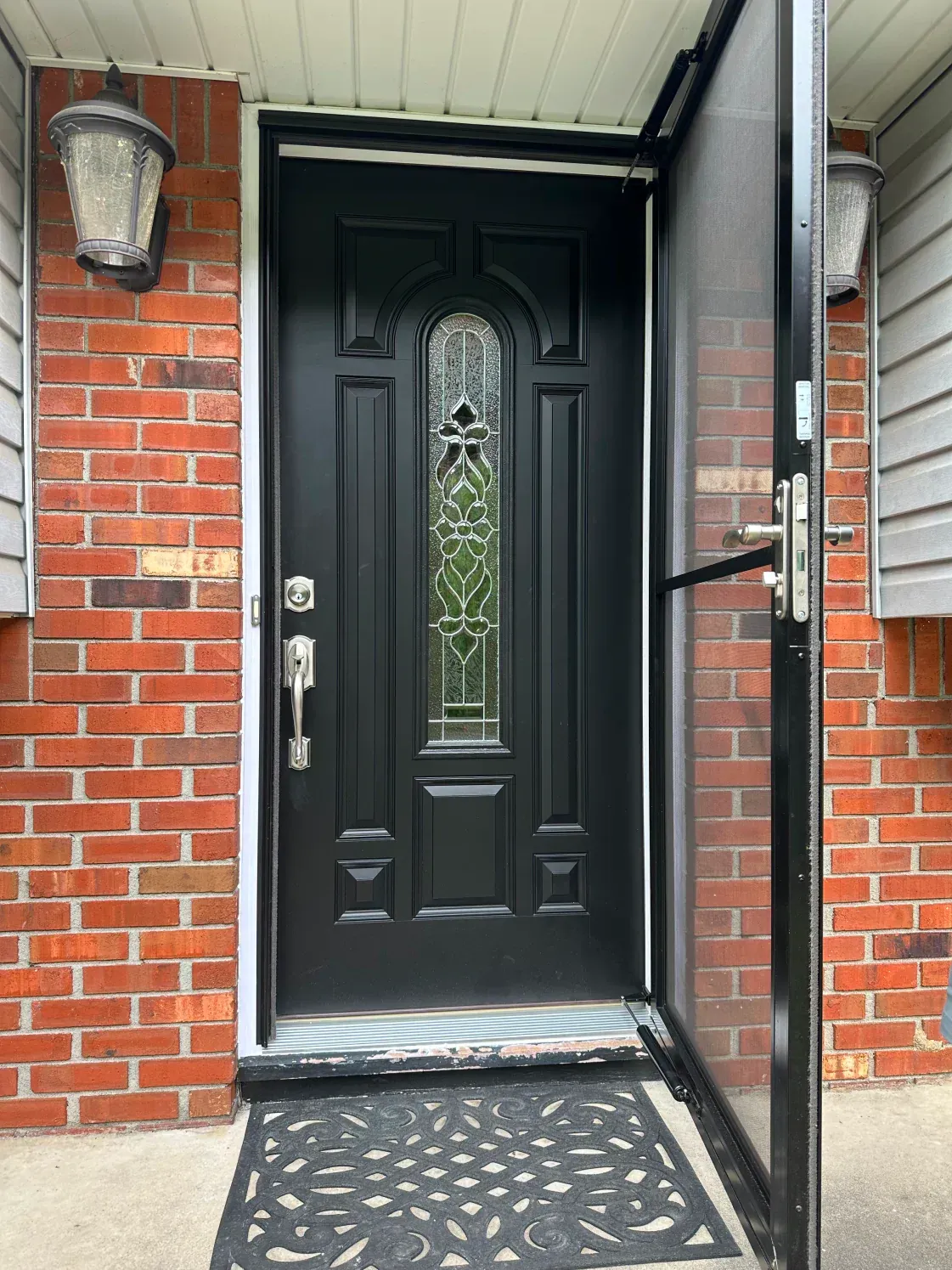 Black front door with decorative glass panel, brick wall, and open screen door.
